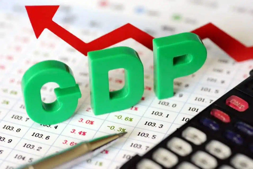 A calculator, a silver pen, and plastic block letters spelling "GDP" sit on a white paper filled with numbers. In the background, a crooked arrow points upward, symbolising economic trends.