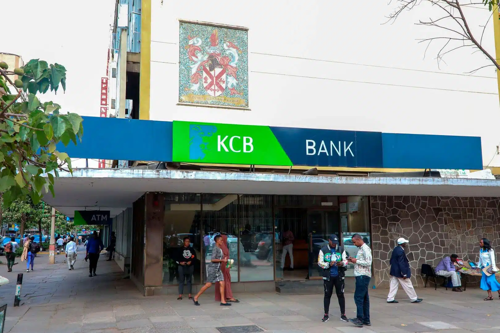 A street-level view of a KCB Bank branch with pedestrians walking past and some standing nearby.