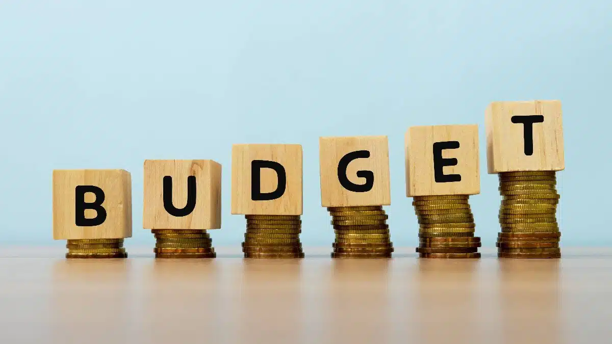 Six stacks of golden coins arranged in ascending order, with wooden cubes on top spelling out 'BUDGET'