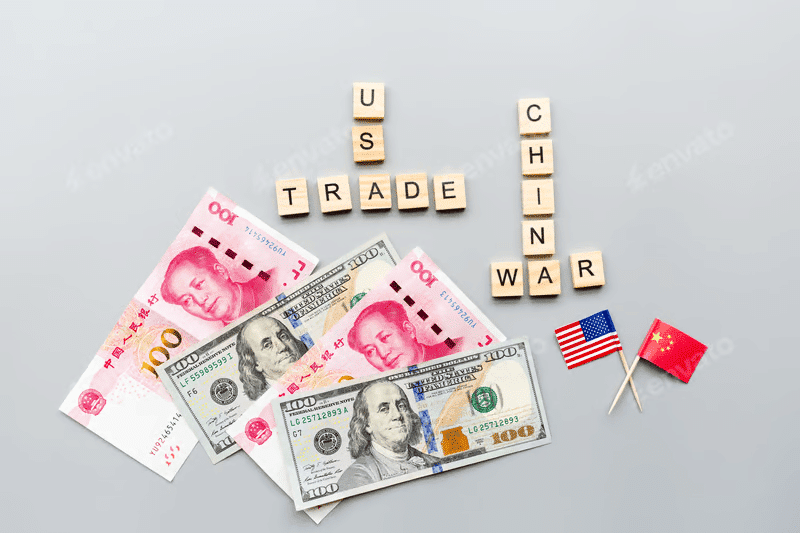 Overlapping U.S. dollar and Chinese yen notes with miniature U.S. and China flags beside them. Above, wooden cubes spell out “U.S.-China Trade War” in bold letters.