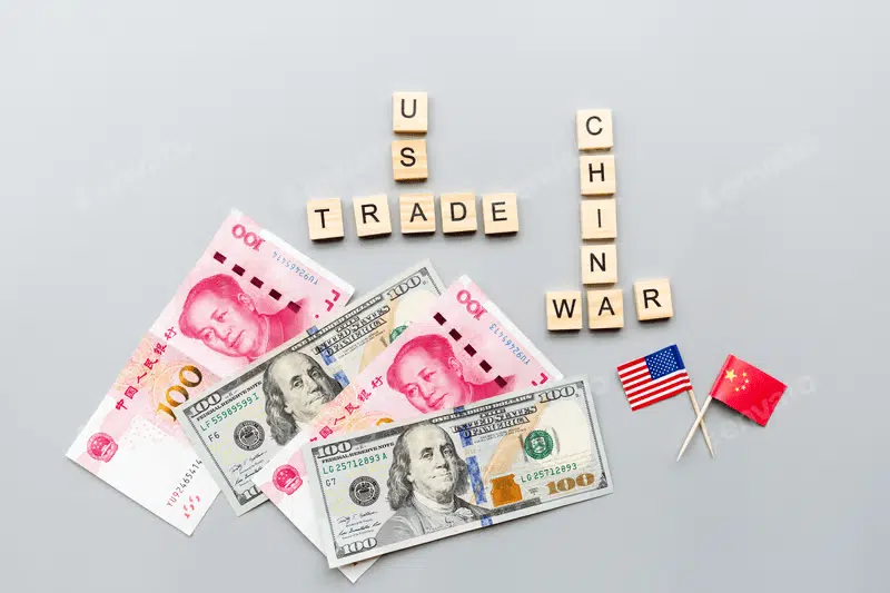 Overlapping U.S. dollar and Chinese yen notes with miniature U.S. and China flags beside them. Above, wooden cubes spell out “U.S.-China Trade War” in bold letters.