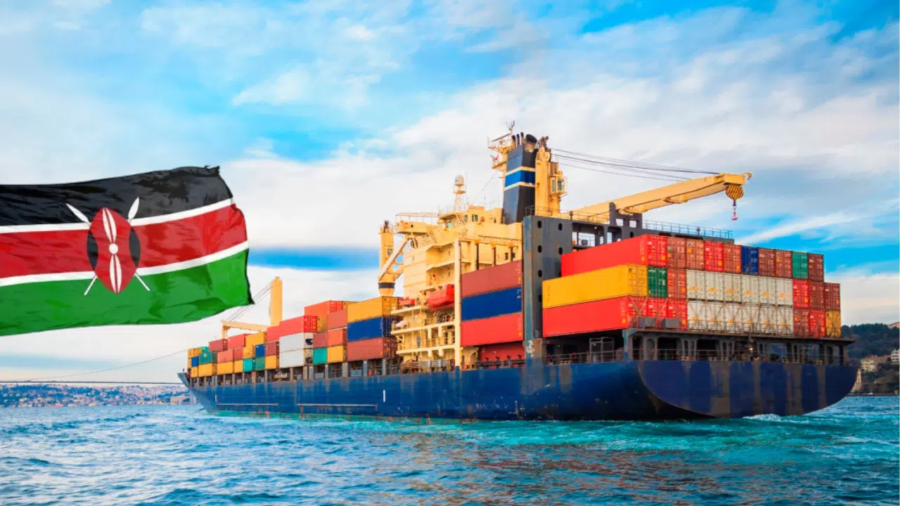 A large cargo ship sailing on turquoise waters, loaded with heavy machinery and multicoloured steel containers. The sky is blue with streaks of white clouds, and a Kenyan flag waves, symbolising trade in Kenya