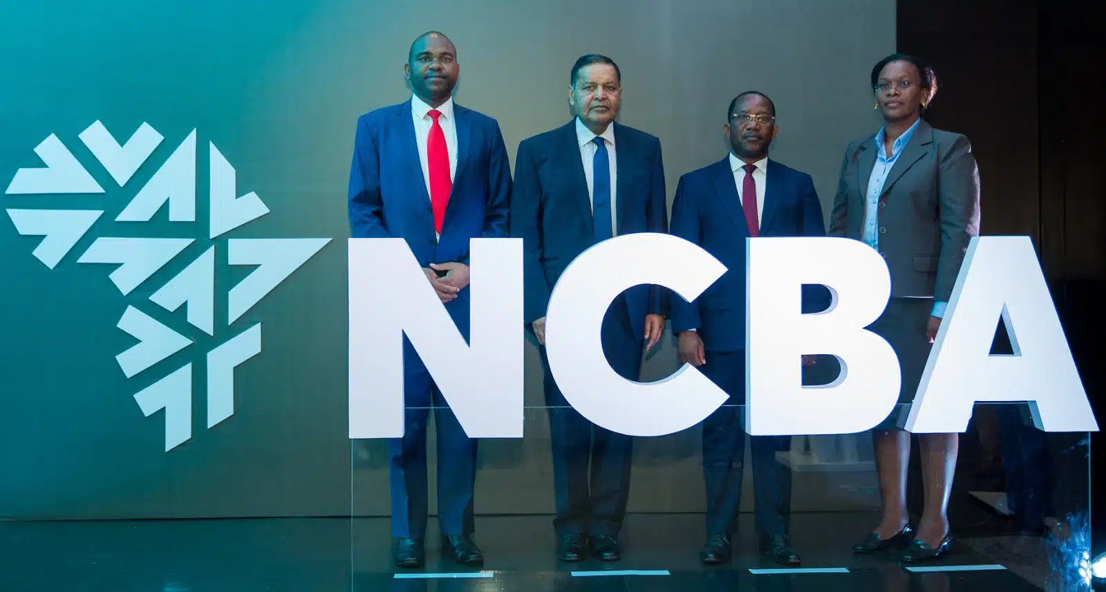 Three men and a woman standing in a line behind a flat horizontal glass holding up large letters that read NCBA during the unveiling of the bank's new logo