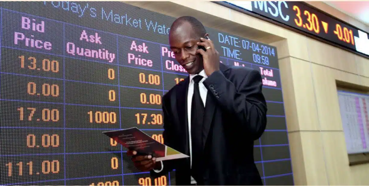 A man talking on the phone while standing in front of an electronic board displaying stock market prices