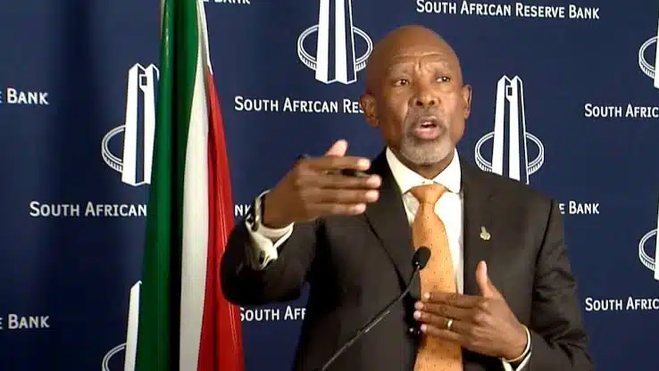 Man in dark suit and orange tie speaking at podium with South African Reserve Bank backdrop and flag visible